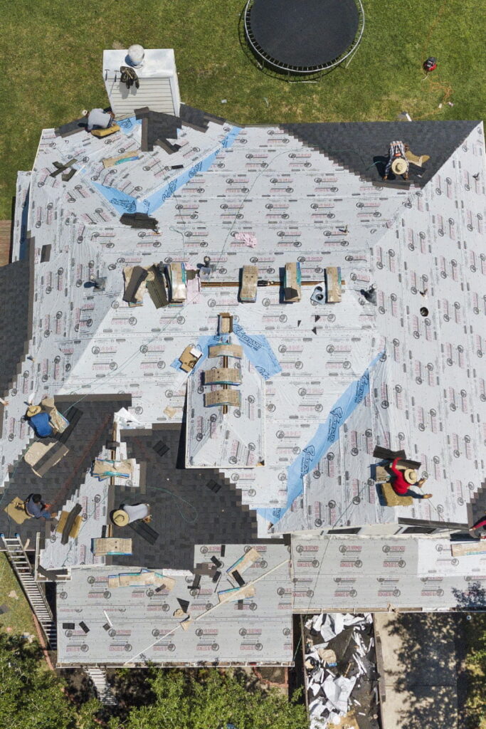 Roofers work on a 2-story house in Houston. Several men replace asphalt shingles and the photography was taken from above using a drone
